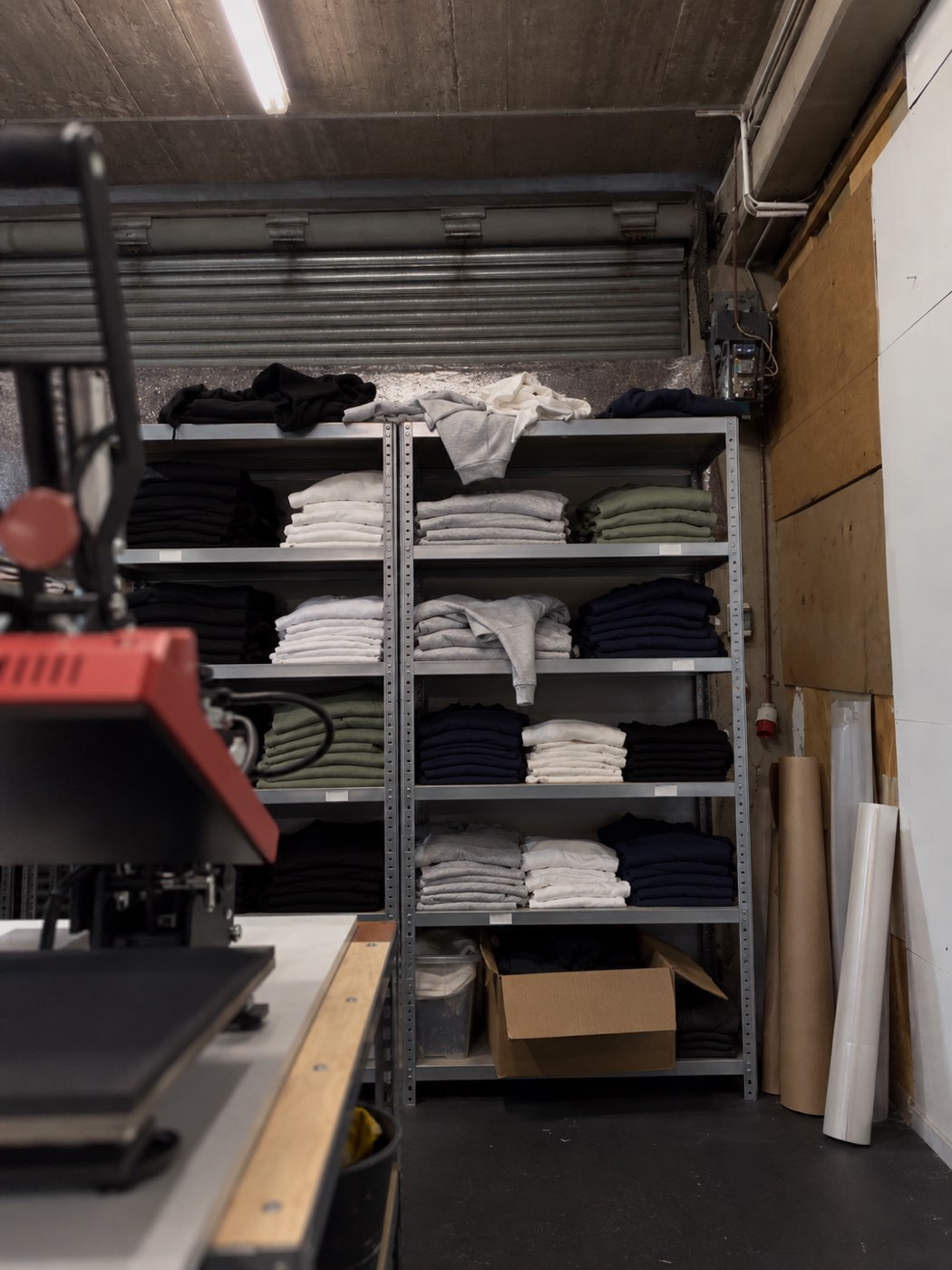 Regal mit gefalteten Rohware-Shirts und Hoodies nach Farben sortiert, davor eine Heißpresse