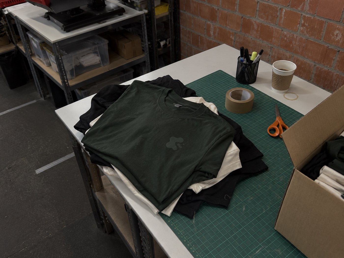 Frisch bedruckte Shirts auf dem Packtisch mit Klebeband, Schere und Versandkarton